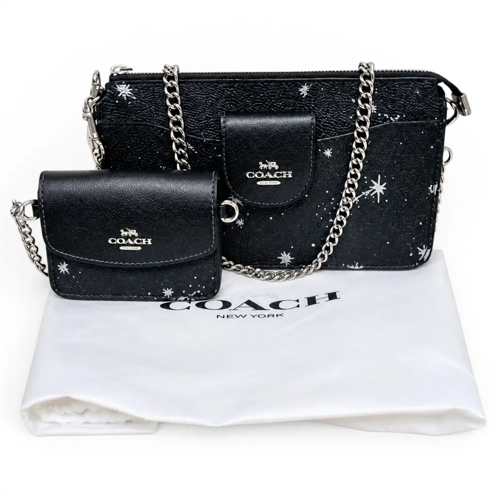 A Coach crossbody bag in black textured leather with silver chain straps and white star print, shown alongside a matching small flap wallet and a white Coach dust bag on a white background. Pre-Owned Coach Black Celestial Print Crossbody Pouch Set – Excellent Condition