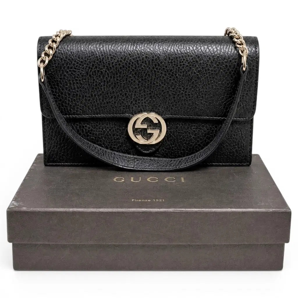 A black pebbled leather Gucci shoulder bag with a gold-tone interlocking GG logo and chain strap, displayed on top of a dark brown Gucci shoebox against a white background. Pre-Owned Gucci Interlocking G Wallet on Chain – Black Leather