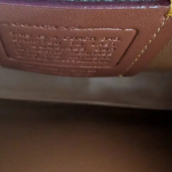 Close-up detail of a brown leather interior label with embossed text, showing part of a tan textured exterior and a light blue glittery trim above. Pre-Owned Coach Tom Wesselmann Rogue Top Handle Bag