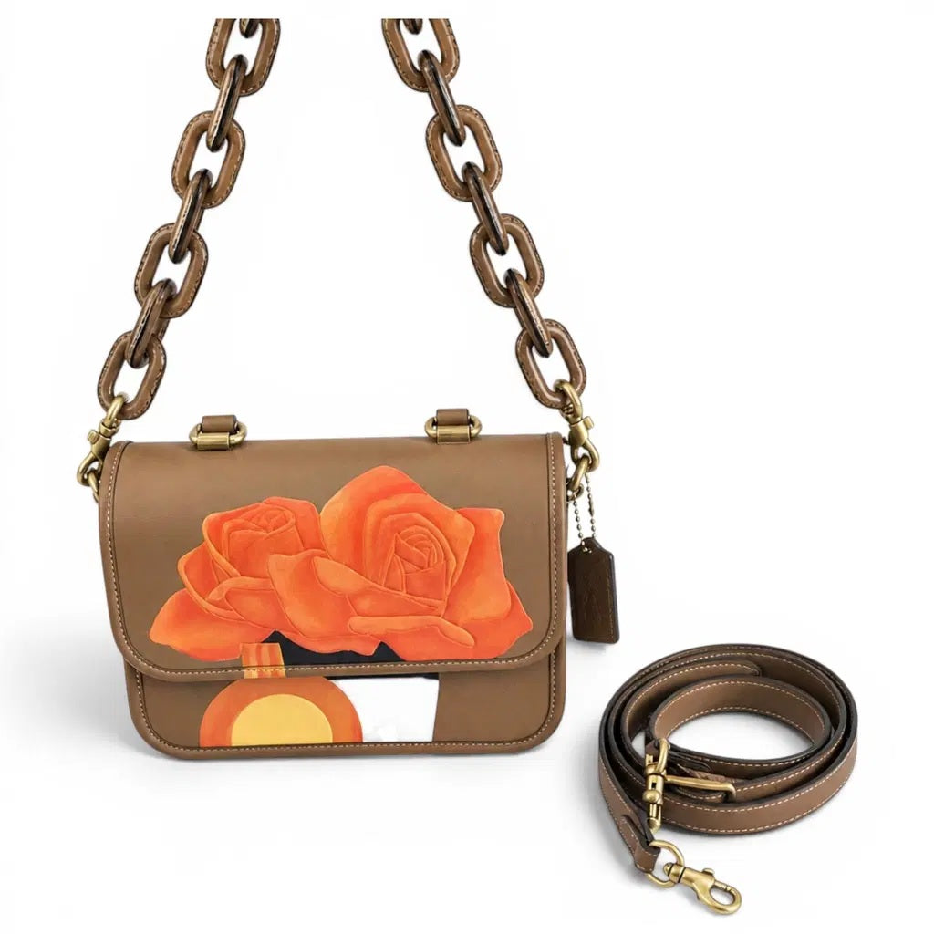 A brown leather shoulder bag with a chain strap and a coiled detachable strap, featuring a large orange rose and sun graphic on the front flap, shown on a white background. Pre-Owned Coach Tom Wesselmann Rogue Top Handle Bag